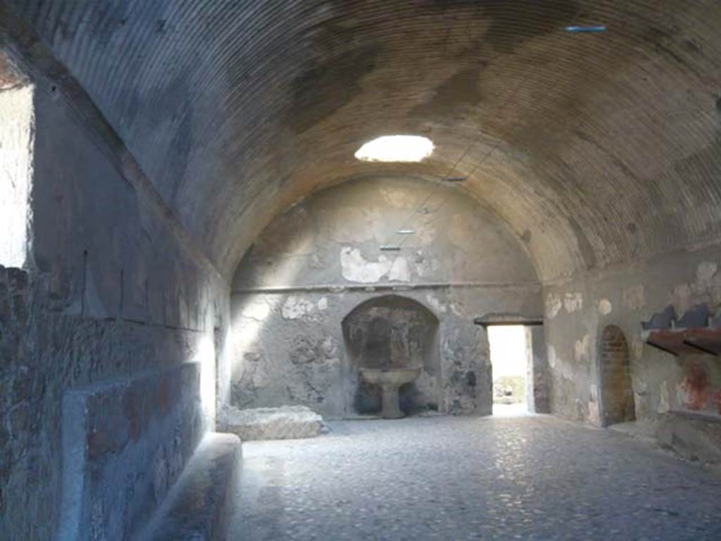 VI.1, Herculaneum. August 2013. Looking north across changing room of men’s baths. Photo courtesy of Buzz Ferebee.
According to Maiuri this was a large room, the best preserved in the men’s section.
Two glass cases in the centre of the room contain the remains of the two skeletons found under the mud-flow on this spot, the first victims discovered in the area of the new excavations; they were probably the custodians of the Thermae who had descended from the upper floor in order to take vain refuge under the robust vault of the tepidarium, when they were submerged under the tide of mud. 
See Maiuri, Amedeo, (1977). Herculaneum. 7th English ed, of Guide books to the Museums Galleries and Monuments of Italy, No.53 (p.37).
(Note: the first edition of this guide book was produced in around 1936, 9 years after the start of the “new excavations”).
