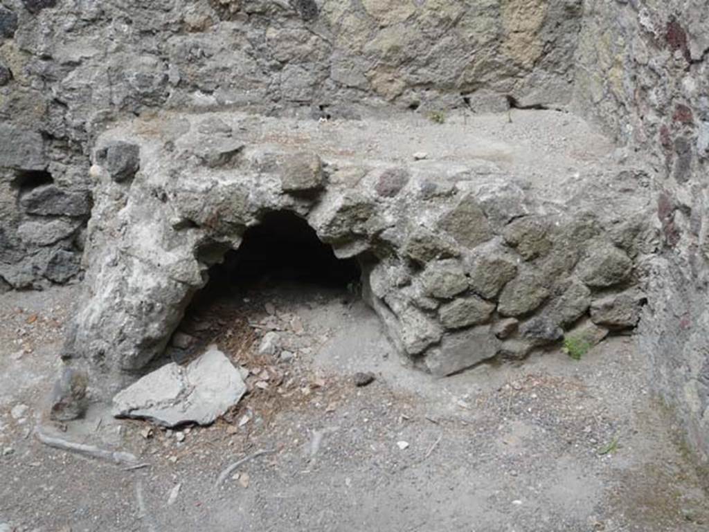 V.35 Herculaneum. August 2013. Bench/hearth in kitchen against west wall. Photo courtesy of Buzz Ferebee.
According to Maiuri –
“Quartiere rustico – Appartato e separato dal resto dell’abitazione era il quartiere rustico.
Vi si accedeva dalla lunga e stretta fauce del lato di nord e si componeva delle parti essenziali: d’una cella penaria (no.7) arieggiata da una finestra, del focolare della cucina allogato nell’angolo di un’area che doveva essere semi-coperta da tettoia (a), della latrina (b) seminascosta da una rampa della scala che saliva alle stanze del piano superiore. Sul muro di contro al focolare vedesi dipinto un serpente, unico avanzo della pittura di un Larario che proteggeva anche qui dall’alto il focolare della casa. Nulla resta delle stanze del piano superiore che doveva estendersi da questo lato, oltreche sull’area del quartiere rustico, sull’esedra no.4 e sul cubicolo no.5.”

(Rustic quarter – the rustic quarter was secluded and separated from the rest of the house. It was accessed from the long and narrow corridor on the north side and consisted of the essential parts: cella penaria (no.7) ventilated by a window, of the kitchen hearth located in the corner of an area that was semi-covered by a roof (a), the latrine (b) half hidden by a flight of stairs leading up to the rooms on the upper floor. On the wall against the hearth, there is a painting of a serpent, the only remnant of the Lararium painting that also protected the hearth of the house from above. Nothing remains of the rooms on the upper floor, which must have extended on this side, as well as over the area of the rustic quarter, to exedra no.4 and cubiculum no.5.)
See Maiuri, A., 1958. Ercolano, I Nuovi Scavi, (1927-1958). Vol.1. Roma: Inst. Poligrafico dello Stato, (p.383).

