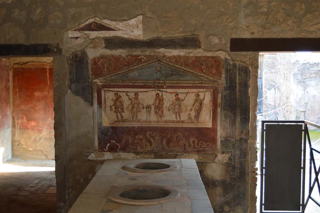 I.8.8 Pompeii. December 2018.Lararium on south wall. Photo courtesy of Aude Durand.
The central figure is the Genius of the household next to a three-legged altar.  
On either side of him are the two Lares. On the left side is Mercury with his money bag.  
On the right side is Bacchus with his panther drinking from a cup held in his right hand.  
Underneath are two serpents approaching a round altar.  
See Fröhlich, T., 1991, Lararien und Fassadenbilder in den Vesuvstädten.  Mainz: von Zabern. (L8: p.252).
