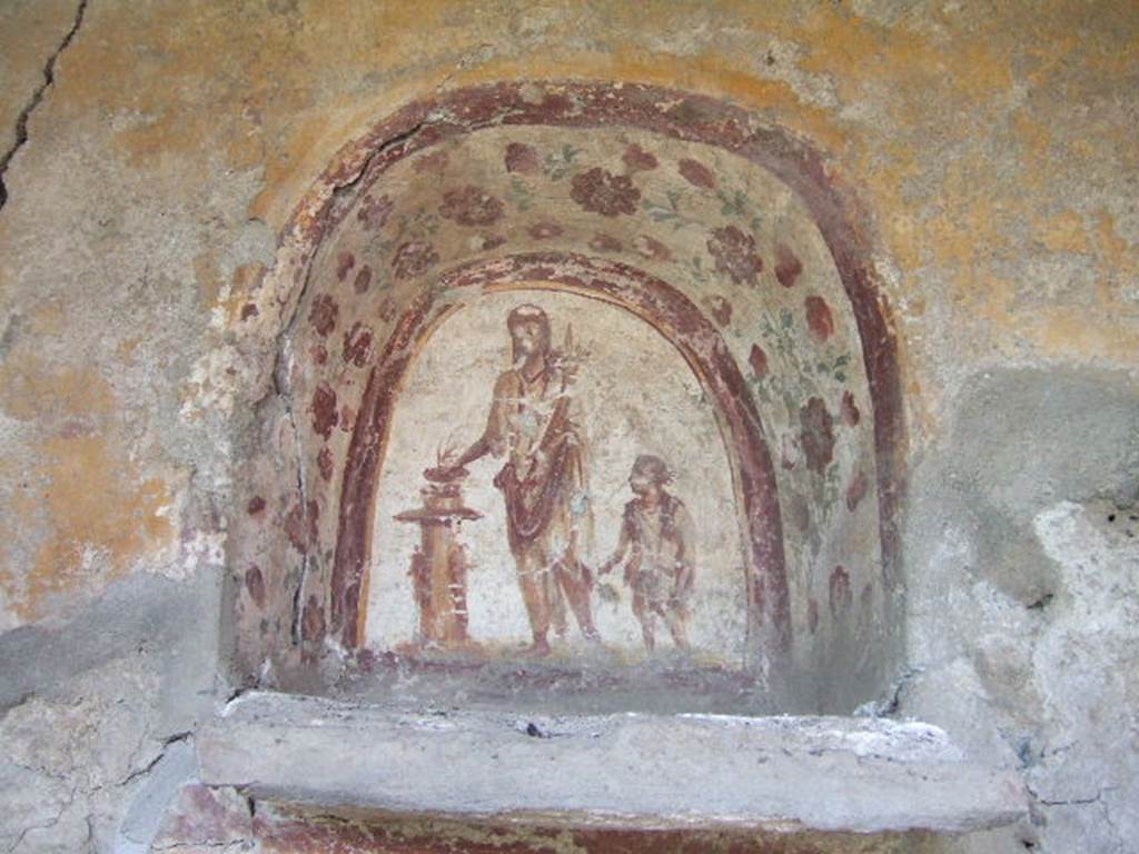 I.17.4 Pompeii.  May 2006.  West wall of peristyle garden.  Lararium niche.
Genius with patera in right hand and cornucopia in the left hand is sacrificing at a round altar.   A small child with a brocca or jug is on the left of the genius. See Bragantini, de Vos, Badoni, 1981. Pitture e Pavimenti di Pompei, Parte 1. Rome: ICCD.  (p.204/5).
