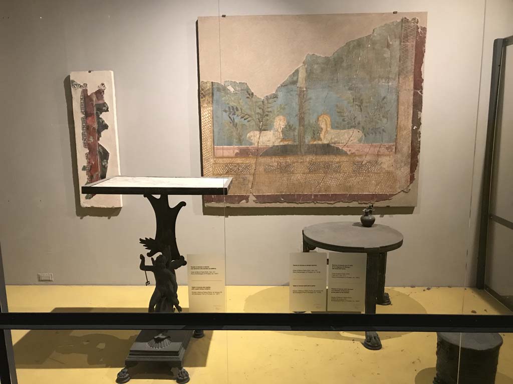 VII.16.17-22 Pompeii. April 2019. On display in Antiquarium, from House of Fabius Rufus. 
Bronze table with cupid riding a dolphin and marble top. PAP inventory number 13371.
Bronze table with lion's feet. PAP inventory number 13108.
On table is a bronze pitcher with face of a follower of Dionysus. PAP inventory number 14072.

Fragment of fresco on the wall at the rear, from VI.17.42, Pompeii. 
Fragment of the wall of the summer triclinium 31 decorated with a pair of facing Sphynxes within a lush garden.
Parco Archeologico di Pompei, inventory number 87228.
Photo courtesy of Rick Bauer.
