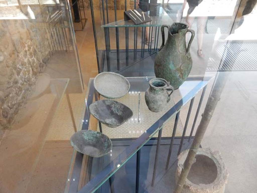 VIII.2.1 Pompeii. May 2018. Finds from excavations conducted in the area since the 1930’s.
The top shelf includes 
A bronze needle. Pompeii inventory number 6312.
Bronze jug with female face at lower joint of the handle. Pompeii inventory number 6319.
Small bronze amphora decorated with theatre masks at the lower joints of the handles. Pompeii inventory number 6321.
Three bronze cups, bronze needle, bronze jug with female face and small bronze amphora decorated with theatre masks. Pompeii inventory numbers 6310A, 6310B, 6323.
All were found in the area to the south of the House of Championnet.
Photo courtesy of Buzz Ferebee.

