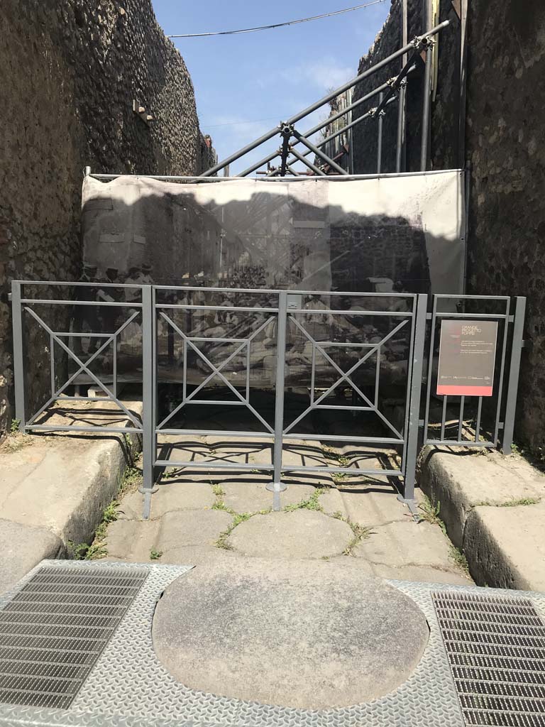Vicolo di Tesmo between IX.1 and IX.7. April 2019. Looking north from Via dell’Abbondanza.
The screen hiding the vicolo shows an enlargement of a photo of the “first workers resting in the shade of the vicolo and posing for their photo.”
See Spinazzola, V. (1953). Pompei, alla luce degli Scavi Nuovi di Via dell’Abbondanza, 1910-1923. (p.31, fig.30)
Note the modernised accessible stepping-stone in the above photo.  Photo courtesy of Rick Bauer.

