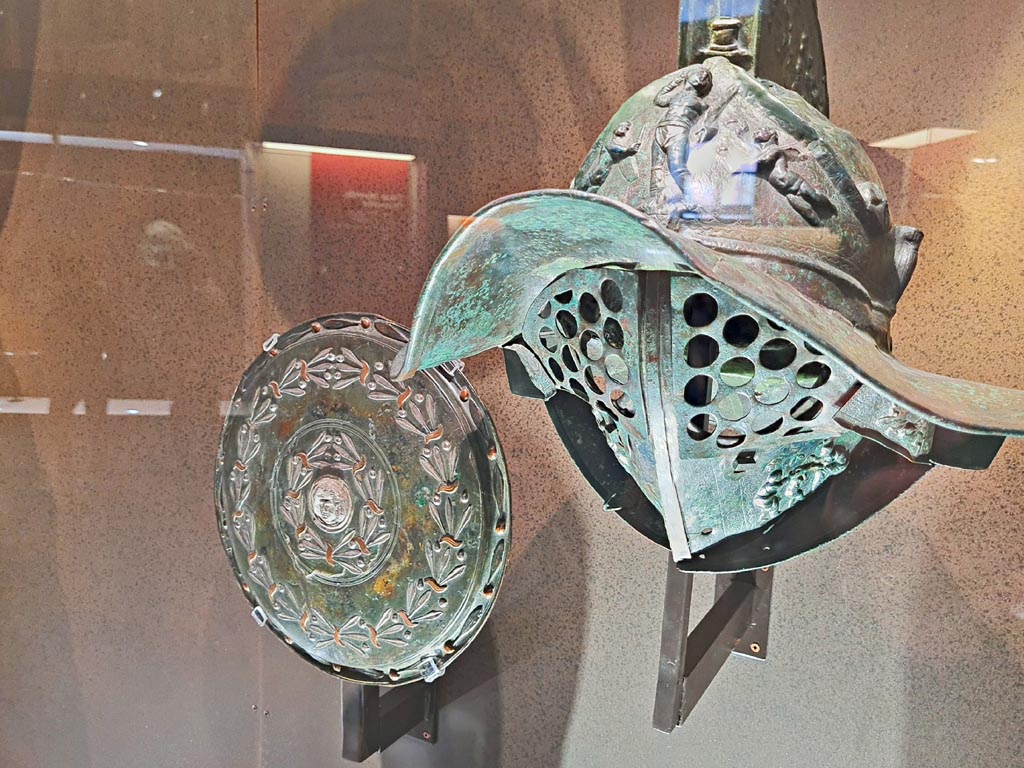 VIII.7.16 Pompeii. Photo taken May 2021, in Naples Archaeological Museum.
On right - Murmillo gladiator’s helmet, showing Gladiator between armed cupids and a herm of Hercules and Bacchanalian scenes on crest inv. 5672.
On left - Bronze, copper and silver shield with laurel branches and gorgeinon, inv. 5669. Found in Gladiators Barracks, 9th January 1768.
Photo courtesy of Giuseppe Ciaramella.