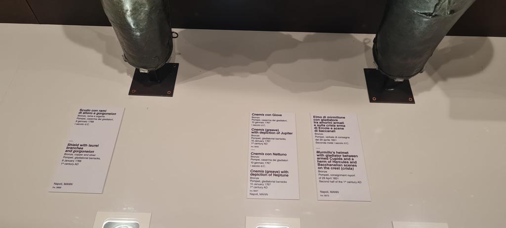 VIII.7.16 Pompeii. Description cards from Naples Archaeological Museum. Photo taken May 2021.
On left – Bronze, copper and silver shield with laurel branches and gorgoneion, inv. 5669.
Centre – Bronze greave with depiction of Jupiter, inv, 5645, bronze greave with depiction of Neptune, inv. 5647.
On right – Bronze Murmillo helmet showing Gladiator between armed cupids and a herm of Hercules and Bacchanalian scenes on crest.
Found in Gladiator’s Barracks, consignment report of 29th April 1851. Photo courtesy of Giuseppe Ciaramella.