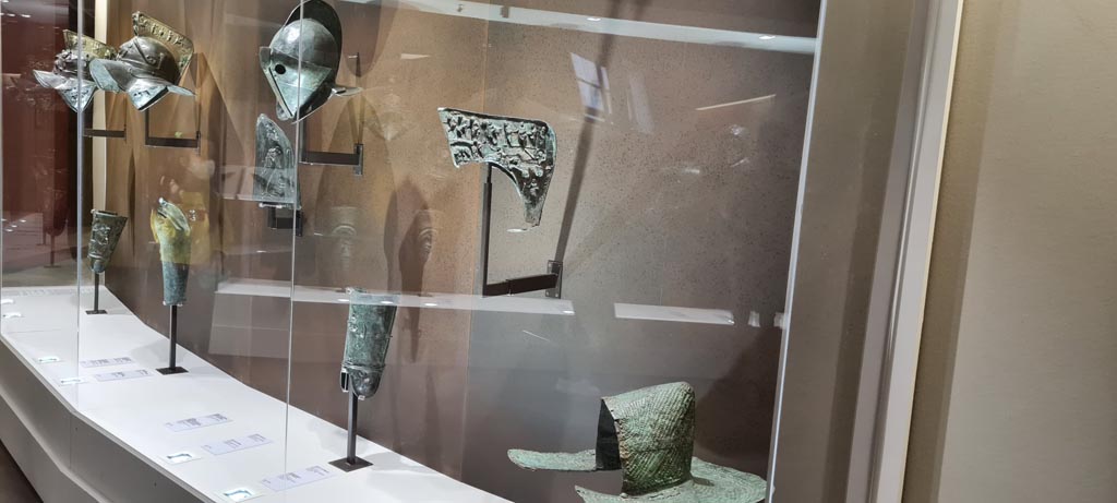 Items in display cabinet in Naples Archaeological Museum. Photo taken May 2021, courtesy of Giuseppe Ciaramella.
On far left –Helmet and greave, inv. 5671 and 5676 (from Pompeii VII.11.6).
Then – helmet with relief of head of Medusa
Then – Secutor’s helmet, inv. 5642.
Then – (from Herculaneum) crest of Murmillo’s helmet with depictions of Mars, Rhea Silvia, she-wolf with Romulus and Remus and deities, found 19th June 1834. Naples Archaeological Museum, inv. 5656.
Lower right - Bronze Pileus (pilos helmet) inv. 5651.