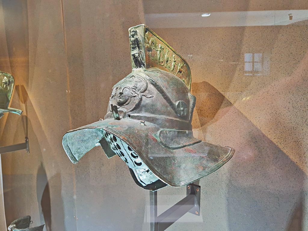 Helmet with relief of head of Medusa. Photo taken May 2021, courtesy of Giuseppe Ciaramella.
Murmillo gladiators carried heavy equipment, including a broadsword and a large, rectangular shield.
The relief on the helmet’s crest depicts griffins being offered drinks by erotes (cupids).
Feathers could be inserted into tubes attached to the sides of the crest.
The head of Medusa, a mythological monster with snakes for hair, can be seen above the visor.
Above this is Mars Ultor, the god of war, in full armour.
