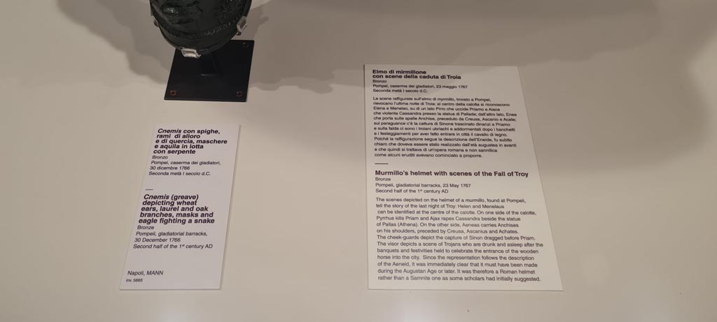 VIII.7.16 Pompeii. Photo taken May 2021, in Naples Archaeological Museum,
Description card –
On left - Bronze greave depicting wheat ears, laurel and oak branches, masks and eagle fighting a snake, inv. 5665.
On right – Murmillo gladiator’s helmet with scenes of capture of Troy, discovered Pompeii on 23rd May 1767 in Gladiator’s Barracks, inv. 5673.
Photo courtesy of Giuseppe Ciaramella.