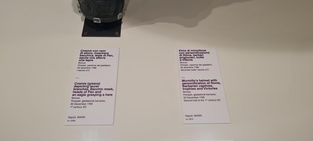 VIII.7.16 Pompeii. Photo taken May 2021, in Naples Archaeological Museum.
Description card –
On left - Bronze greave depicting laurel branches, Bacchic masks, heads of Pan and an eagle clasping a hare, inv. 5668.
On right - Murmillo gladiator’s helmet, with personification of Rome, Barbarian captives, trophies and Victories, inv. 5674.
Found in Gladiator’s Barracks, 20th December 1766. Photo courtesy of Giuseppe Ciaramella.