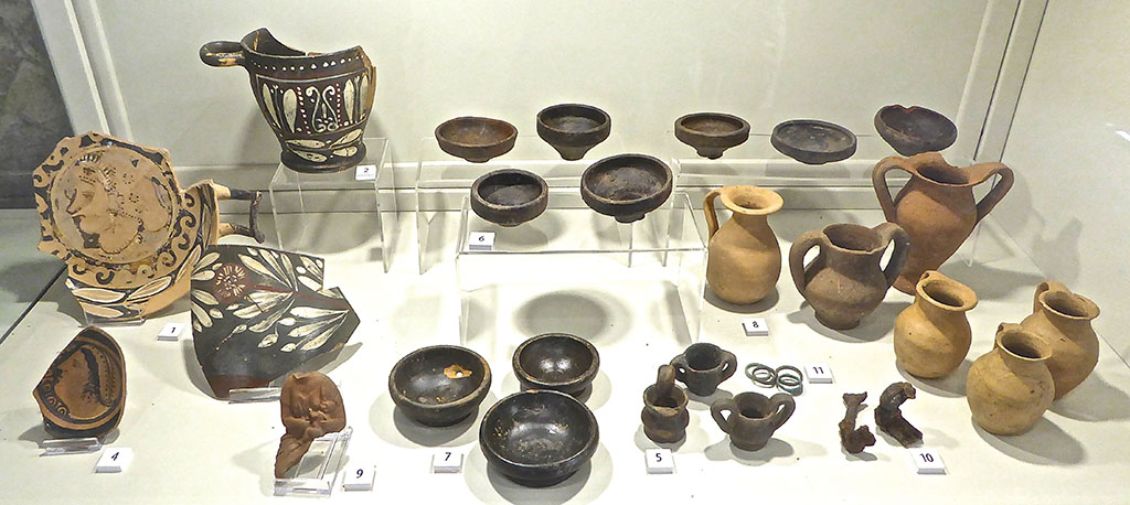 Santuario extraurbano del Fondo Iozzino. June 2017. Finds from site. Now in PAP deposits.
Photo courtesy of Michael Binns.
The exhibition information card identifies these as:
1: Red figure kylix with woman’s head, 4th century BC
2: Campanian painted skyphos, second half of 4th century BC
3: Rim of painted Campanian bowl, second half of 4th century BC
4: Base of Campanian bowl with female head, second half of 4th century BC
5: Miniature kraters, 4th to 2nd century BC
6: Miniature plates, 3rd to 2nd century BC
7: Black gloss cups, 3rd century BC
8: Coarse ware beakers, 4th to 2nd century BC
9: Terracotta figurine of woman nursing a child, 4th to 3rd century BC
10: Votive iron keys, Hellenistic period
11: Bronze rings, Hellenistic period
