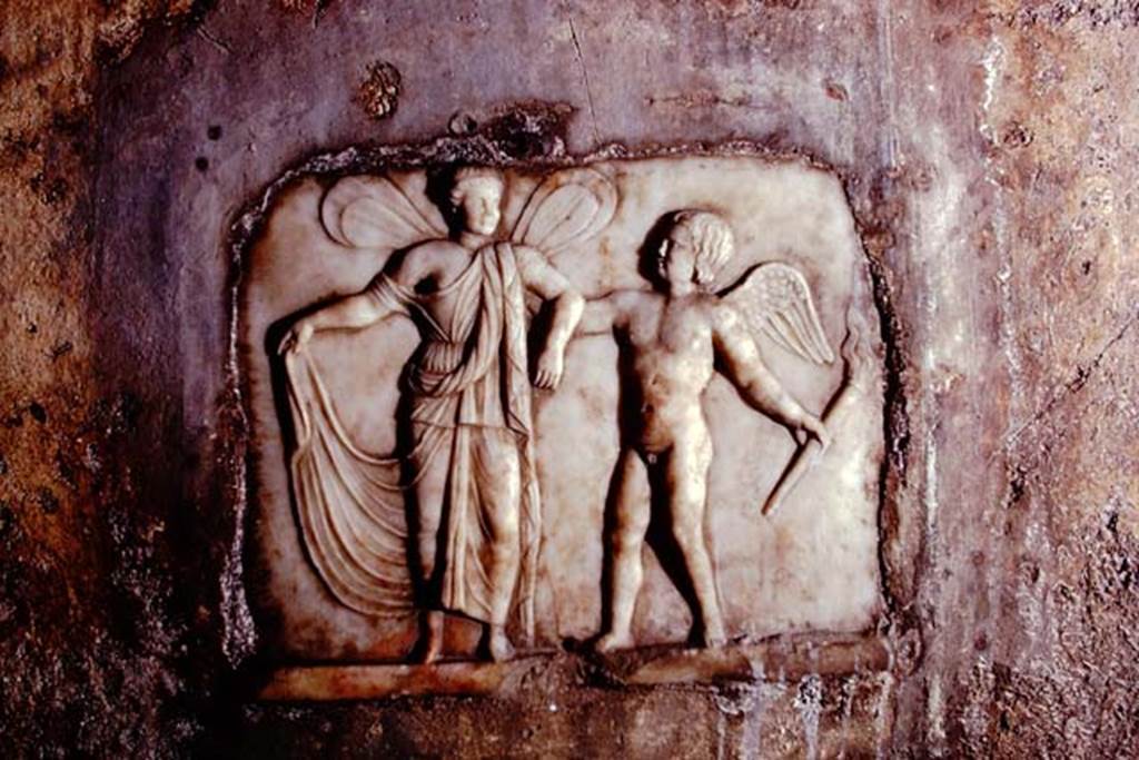 Santa Maria Capua Vetere, Mithraeum. 1974. South wall, white marble relief of Eros and Psyche.
Photo by Stanley A. Jashemski.
Source: The Wilhelmina and Stanley A. Jashemski archive in the University of Maryland Library, Special Collections (See collection page) and made available under the Creative Commons Attribution-Non-Commercial License v.4. See Licence and use details.
J74f0582
The white marble relief (H. 0.32 Br. 0.39) was found in a recess in the southern wall above the water-basin of the podium.
The nude winged child Amor seizes the arm of the bigger Psyche, also winged, to lead her away in the light of his torch. The girl lifts a part of her diaphanous dress. The whole is set in a red frame.
See Vermaseren M. J., 1971. Corpus Inscriptionum et Monumentorum Religionis Mithriacae, p. 108, no. 186, fig. 56.
See Minto A., in Notizie degli Scavi di Antichità, 1924, p. 367; fig. 9.