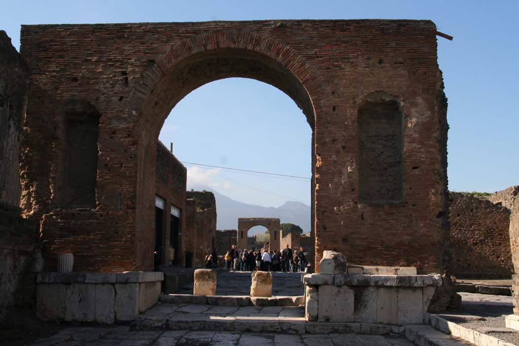 Arch at north-east end of the Forum. April 2013.  
South side, looking north through arch onto Via del Foro, and towards Arch at south end of Via di Mercurio, with Vesuvius in the distance. 
Photo courtesy of Klaus Heese. 
