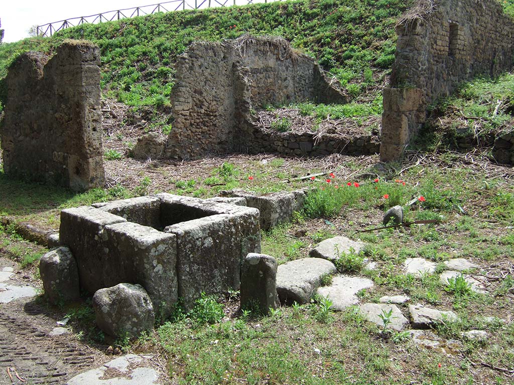 Via di Nola. May 2006. Looking north-east across street fountain on corner by III.11.1.
According to Eschebach, this fountain only had a basin and a fragment of the rear pilaster and no relief.
See Eschebach, H., 1983. Pompeii, Herculaneum, Stabiae; Bollettino dell’Associazione Internazionale Amici di Pompei 1, p. 12, Kat. Nr. 10. 
