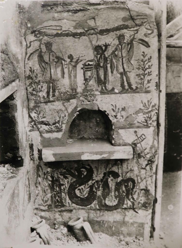I.8.14 Pompeii. 1938. 
Lararium in north-east corner of courtyard 7, near to the kitchen (10), no longer conserved.
See Fröhlich, T., 1991. Lararien und Fassadenbilder in den Vesuvstädten. Mainz: von Zabern, See Fröhlich, T., 1991. Lararien und Fassadenbilder in den Vesuvstädten. Mainz: von Zabern, (p. 254, L10, Taf. 26,2).
Above the niche is the painting showing two large Lares, a smaller togated Genius with a cornucopia and sacrificing on a round altar while on the other side of the altar a tibicen is playing the double flutes.
At the sides of the niche are two birds in shrubs.
Below the niche is a serpent approaching an altar.
See Giacobello, F., 2008. Larari Pompeiani: Iconografia e culto dei Lari in ambito domestico.  Milano: LED Edizioni. (p.141, no.13).
