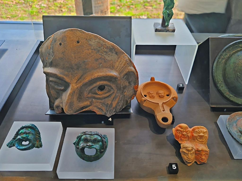 I.9.13 Pompeii. March 2024. No.5 – Plaque with theatre masks, (Ist century BC – Ist century AD). Photo courtesy of Giuseppe Ciaramella.
No.3, lower left – Bronze wall sconces with theatre masks, provenance not known, but from Pompeii. (Ist century AD).
No.4 – Terracotta theatre masks Pompeii – provenance not known, but from Pompeii. (Ist century AD). 
No.6 (number hidden, but beneath No.7) – Oil lamp with theatre masks, from I.9.9.
On display in exhibition in Palaestra entitled – “L’altra Pompei, vite comuni all’ombra del Vesuvio”.
