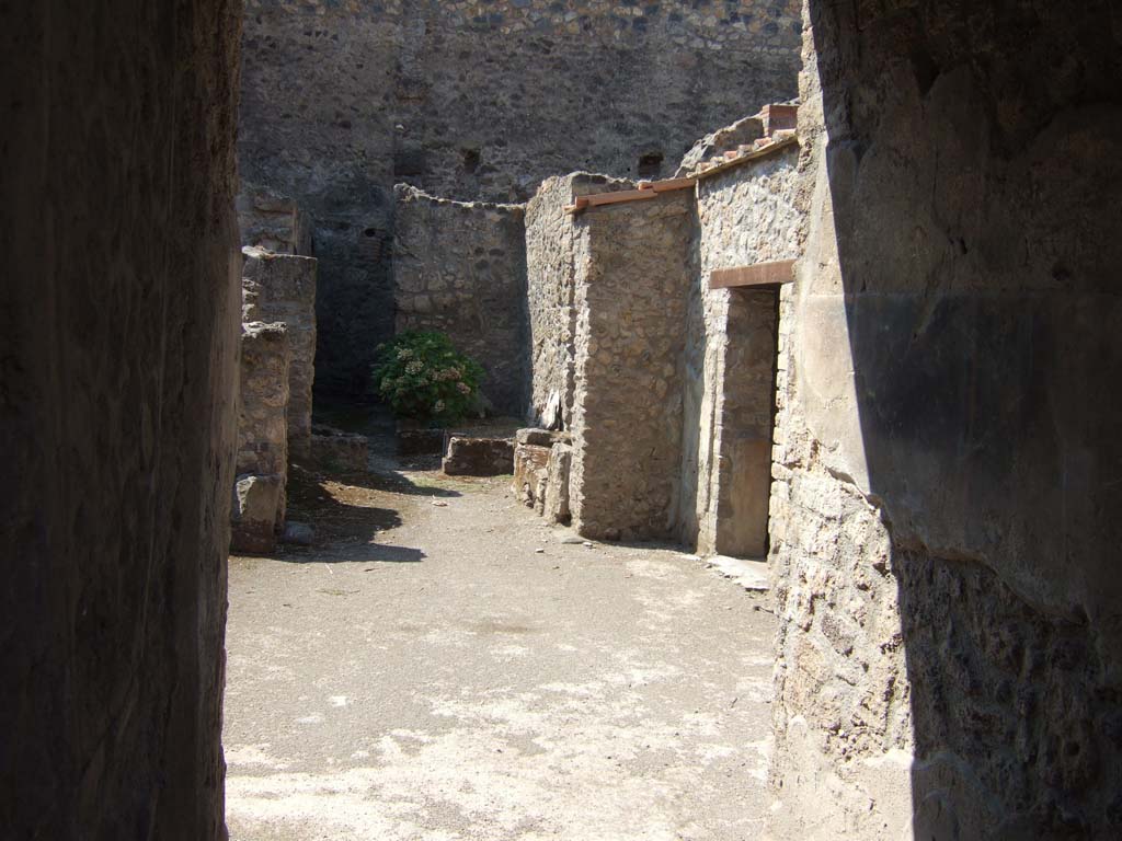 I.10.18 Pompeii. September 2005. Looking north-west from entrance fauces.
On the right is the doorway to Room 5, followed by room 6.
“Along the north wall, next to the wall shared with I.10.1, opened successively, the small rectangular ala (no.4) in the form of an exedra finely decorated with painted panels with a black background and white and yellow-gold threads, and then another small room used as an apotheca (no.5).
The decoration of the ala/exedra was of the III Style, with a black painted zoccolo with white vertical lines joined by a thin red band, finished with a narrow green band. The background of the wall was divided into red panels by thin white lines, and with white lines of panels and finished at the top with a green band decorated with floral elements.
The upper part of the wall, also with a red background, presented the motif of a slender wooden architecture consisting of an aedicula with two smaller side aediculae, from which hung suspended light garlands. The flooring was of lavapesto.
The following small room (5), which we will call an apotheca, had a light yellow zoccolo and the upper part of the wall was left with rough plaster.”
The flooring was of concrete (calcestruzzo).
