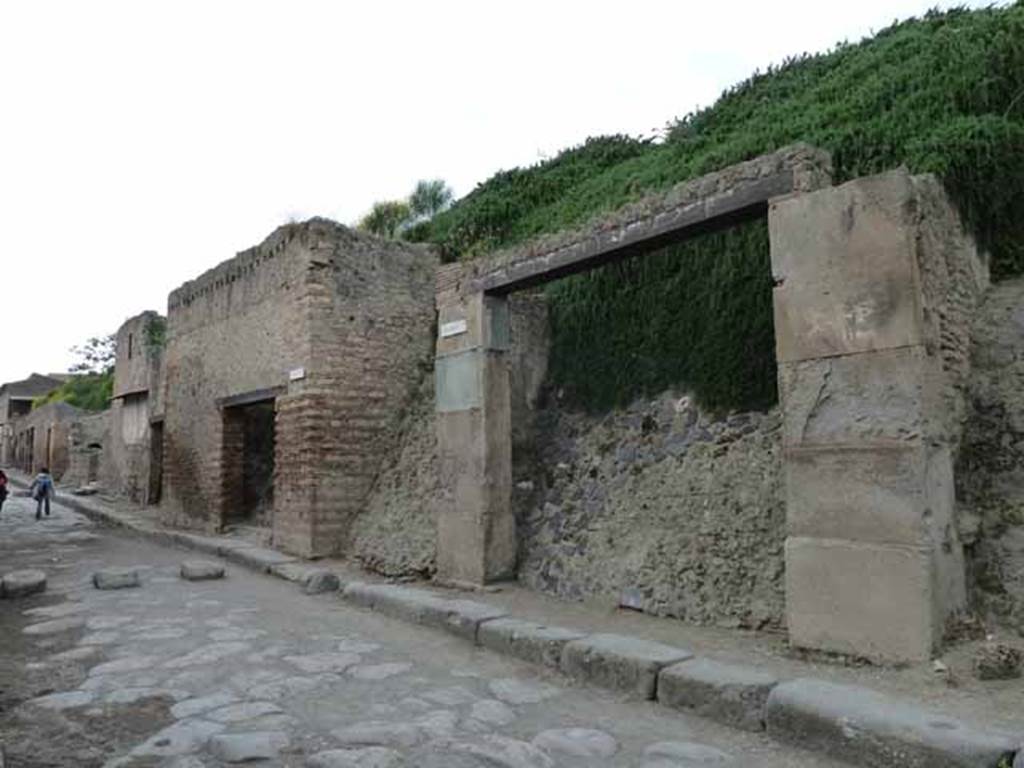 III.3.1 Pompeii. May 2010.  Entrance on Via dell’Abbondanza, looking west past unexcavated roadway to insula III.2.  Originally when excavated in 1914, this doorway was found with carbonised architrave and part of carbonised doorpost.
These were reinstated under glass, and left in situ, after consolidation of the doorway.   See Notizie degli Scavi, 1914, (p.255)
There was also a balcony leaning out over the street, which was reconstituted from its elements,as well as the remains of a stone window frame, to the right (east end).
