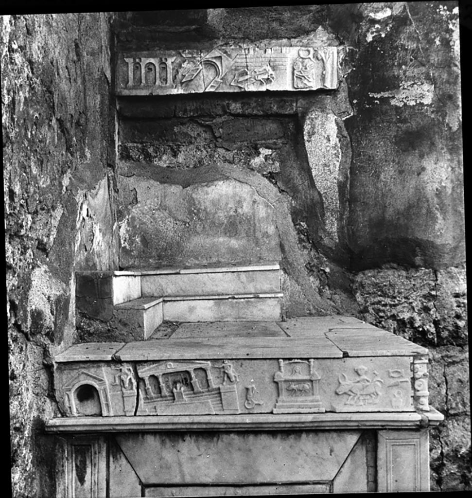 V.1.26 Pompeii. Photo by Nash, Fototeca Unione. Detail of marble lararium in north-west corner of atrium.
The upper marble relief shows scenes of the earthquake of AD62 possibly in Porta Vesuvio area.
It appears to show the castellum aquae, the collapsed Porta Vesuvio, city walls and a collapsed two horse cart/chariot and an altar with offerings and a wreath above.
The lower marble relief shows scenes of the earthquake of AD62 possibly in the Forum.
Used with the permission of the Institute of Archaeology, University of Oxford. File name instarchbx202im 077. Source ID. 44536.
See photo on University of Oxford HEIR database
The upper marble relief was stolen in 1975.

