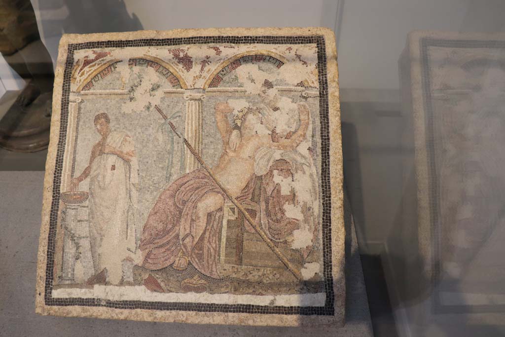 V.6.7 Pompeii. February 2021. Mosaic on display in Pompeii Antiquarium.
Central mosaic picture that adorned a fountain in a house on Via del Vesuvio, in front of which there was a statuette of a rabbit.
Photo courtesy of Fabien Bièvre-Perrin (CC BY-NC-SA).
According to Kuivalainen, this was the centre mosaic below a niche with columns and a pediment. 
The mosaic consists of 3 figures in an architectural setting against a light blue background and white floor area.
On the left stands a man holding his right hand over and looking down on a brownish basin on top of a low separate stand, a white column, entwined with a garland.
The main figures are a couple loosely embracing each other, a reclining drunken youth looking upwards towards a female figure, seated behind, higher up on the podium, looking down at him.
They are without doubt Bacchus and Ariadne.
See Kuivalainen, I., 2021. The Portrayal of Pompeian Bacchus. Commentationes Humanarum Litterarum 140. Helsinki: Finnish Society of Sciences and Letters, D16: p. 138-9.
