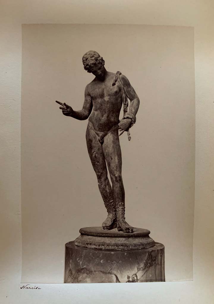 VII.12.17.21 Pompeii. Photograph by M. Amodio, from an album dated April 1878.
Statuette of Narcissus also sometimes described as Dionysus or Pan. Photo courtesy of Rick Bauer.
According to Kuivalainen, this statue (now in Naples Archaeological Museum, inv. no: 5003), had fallen from the upper floor.
The condition is good, but the original base has been replaced, and the panther is lost.
He comments  
The males apparel and the position of the right-hand fingers identifies the bronze as Bacchus dangling a bunch of grapes above a panther.
The iconographic formula is also known from wall paintings in Pompeii.
See Kuivalainen, I., 2021. The Portrayal of Pompeian Bacchus. Commentationes Humanarum Litterarum 140. Helsinki: Finnish Society of Sciences and Letters, (p.219, H14.)
.
