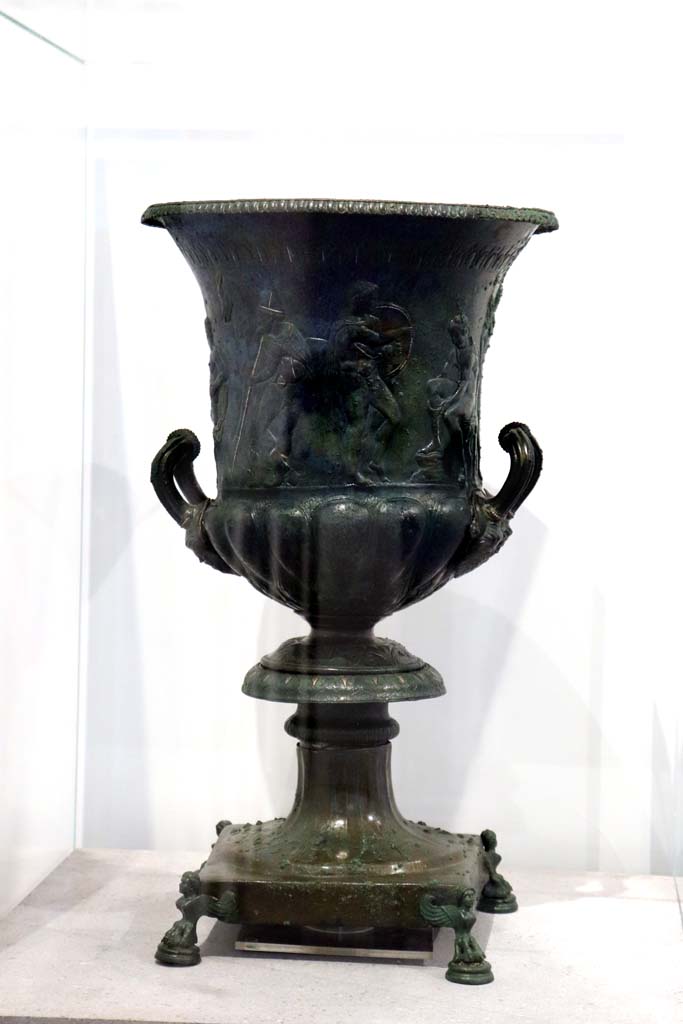 VIII.1.4 Pompeii. February 2021. 
Bronze krater with scenes in relief, found in the triclinium at IX.13.1-3, on display in Antiquarium.
Photo courtesy of Fabien Bivre-Perrin (CC BY-NC-SA).
According to the description card in the Antiquarium -
The bronze krater was decorated with eight armed male figures, six standing and two sitting. 
One of the seated figures is tying his shoelaces.
A young man stands in front of him holding a shield, with a bearded figure holding a staff behind.
Young and naked, the second seated figure caresses an animal with one hand and holds a sword by the hilt in the other.
(See IX.13.1-3 for more photos).
