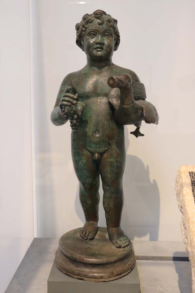 VIII.1.4 Pompeii. February 2021. One of the two bronze statuettes, (as described on P.107 above). 
Bronze statuette of young boy holding a goose and a bunch of grapes, from the peristyle of VI.15.1 (the House of Vettii).   
Photo courtesy of Fabien Bivre-Perrin (CC BY-NC-SA).
