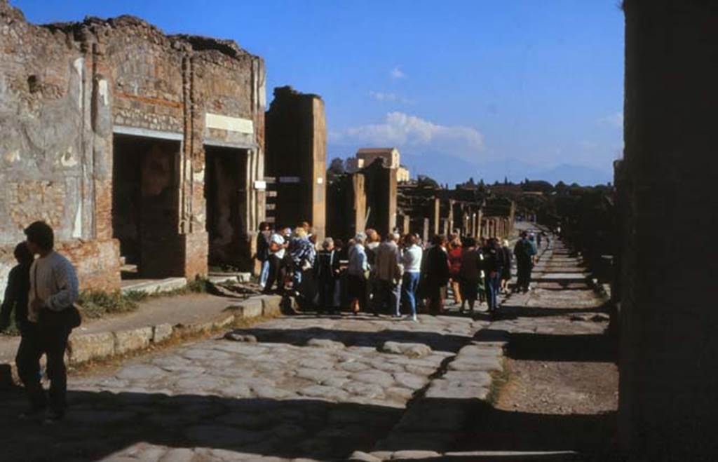 Via dell’ Abbondanza, Pompeii. February 1988.
Looking east, with VII.9.68/67 doorways on left, behind fountain surrounded by crowd of tourists.
Photo by Joachime Méric courtesy of Jean-Jacques Méric.
